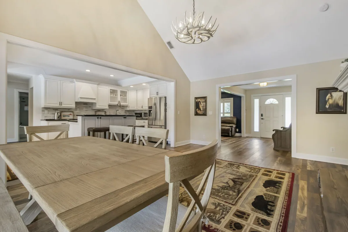 Dining area with a farmhouse table and chairs opening onto a white kitchen under a vaulted ceiling