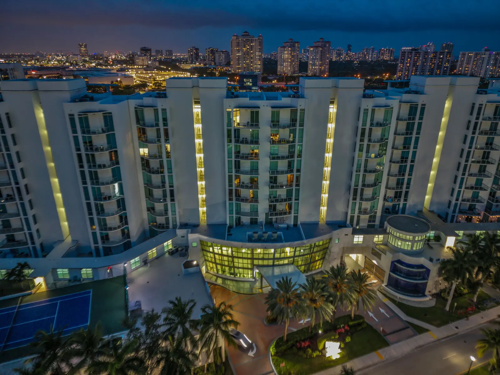 Twilight aerial of a lit Aventura condo entrance and tennis courts